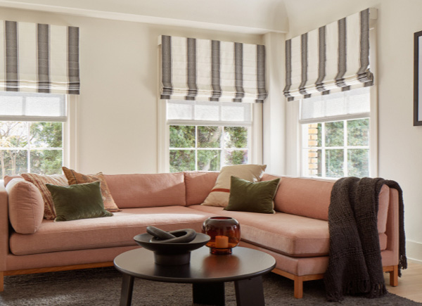 Large windows with Cascade Roman Shades hung over Solar Shades in a living room with dark colored decor and a pink couch