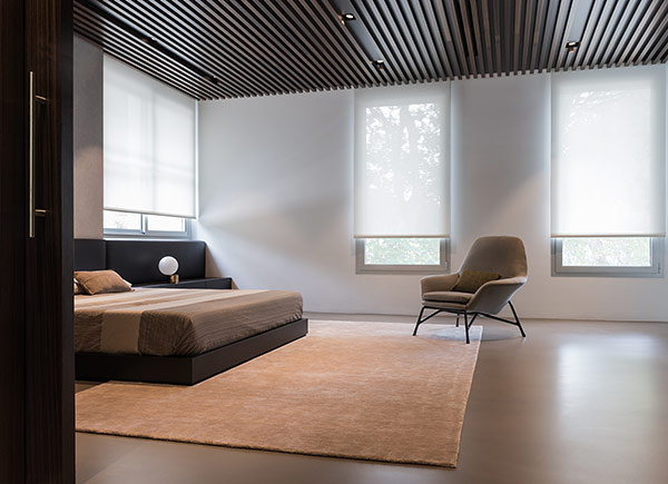 Windows featuring roller shades in 5 percent beige in a bedroom with a low bed and a grey chair over a neutral rug