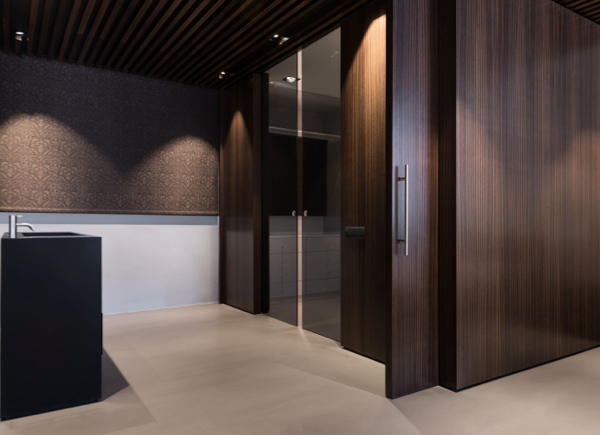 A high set window featuring roller shades in a Bathroom with dark wood walls, black glass shower doors and a black sink