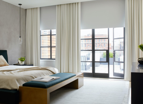 A patio door and large window featuring blackout roller shades layered under drapery in a bedroom with a wooden bench