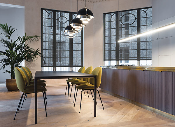 Two large windows featuring roller shades in a kitchen with a white wall of cabinets and large dark table with yellow chairs