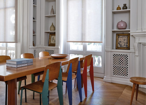 Tall windows featuring roller shades in a Dining Room with built in shelves and a wooden table & chairs with colorful accents