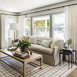 Large windows featuring Tailored Pleat Drapery in a cottage living room with grey couch, brown table and patterned rug