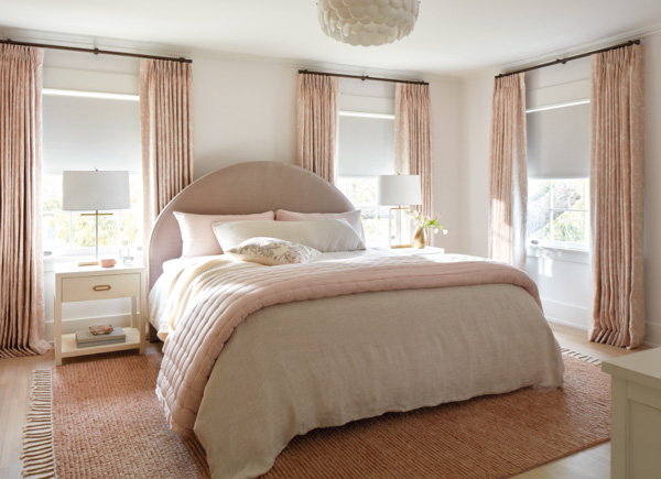 Three windows featuring blackout roller shades layered under tailored pleat drapery in a bedroom with pink and white decor