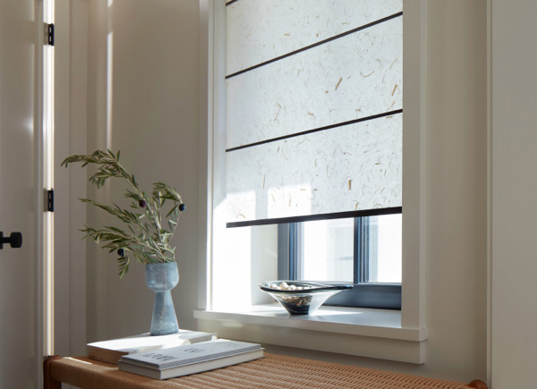 A Zen Roller Shade made of Papyrus in Natural softens the light in a small room with dried leaves in a vase on a desk