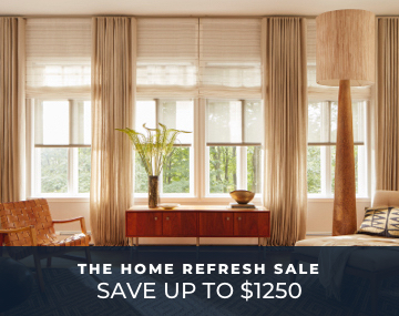 Tall windows featuring roller shades over roman shades and drapery in a living room with chairs and a table with promo text
